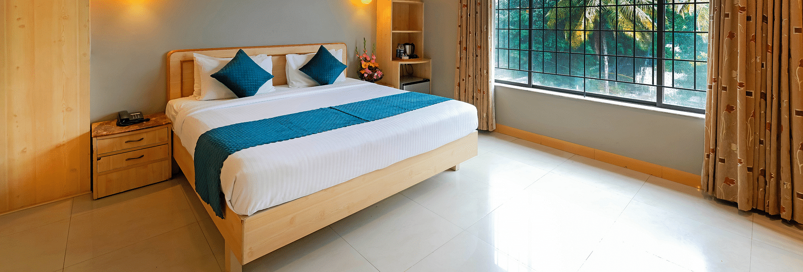 An image of a hotel room in Bengaluru featuring a queen size bed, a night stand, and sunlight from the expansive windows - Cross Roads Inn, Bengaluru