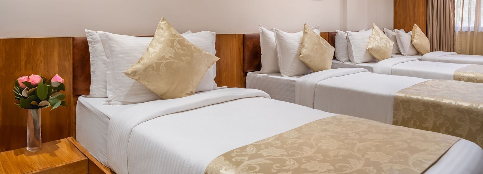Three single beds with white linens and decorative pillows line a hotel room with framed artwork and a window - Lords Eco Inn Ambaji.