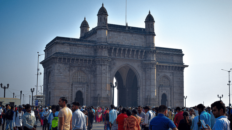 Full view of the Gateway Of India historic monument on a bright sunny afternoon with huge number of pedestrians in the foreground.