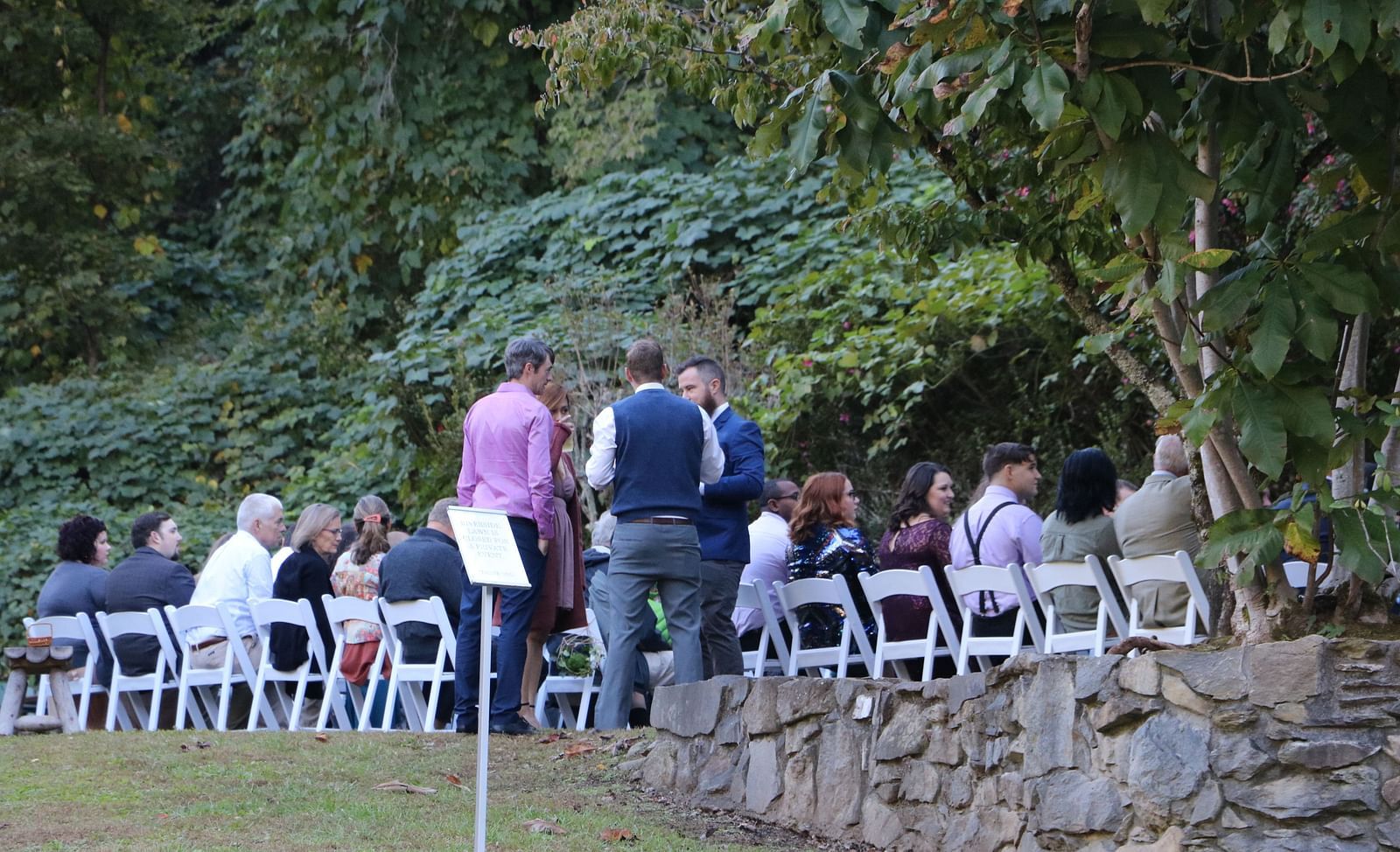 guests gathered outdoors seated on white folding chairs near a stone wall surrounded by lush greenery - Historic Tapoco Lodge
