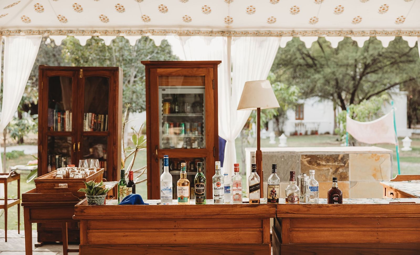 A bar setup with bottles, cabinets, and a lamp under a tent, with trees visible in the background- Shahpura Bagh.
