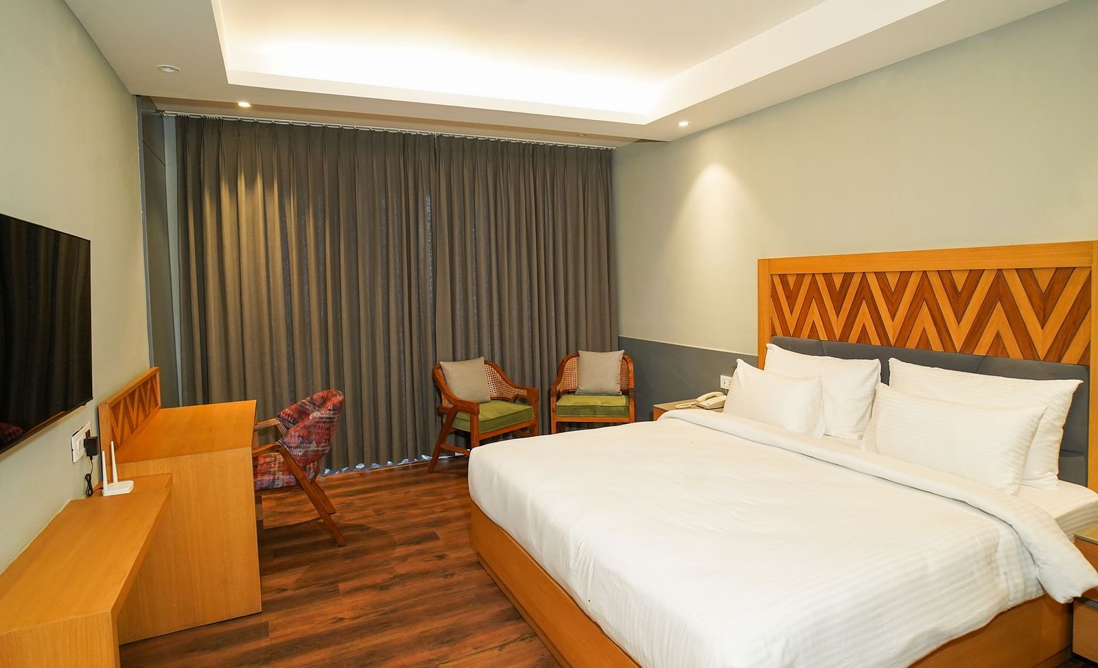 A brightly lit hotel room showing a neatly made bed with white linens and a view towards the window with drawn curtains| Deluxe Room | Sukama Resort & Spa, Nainital