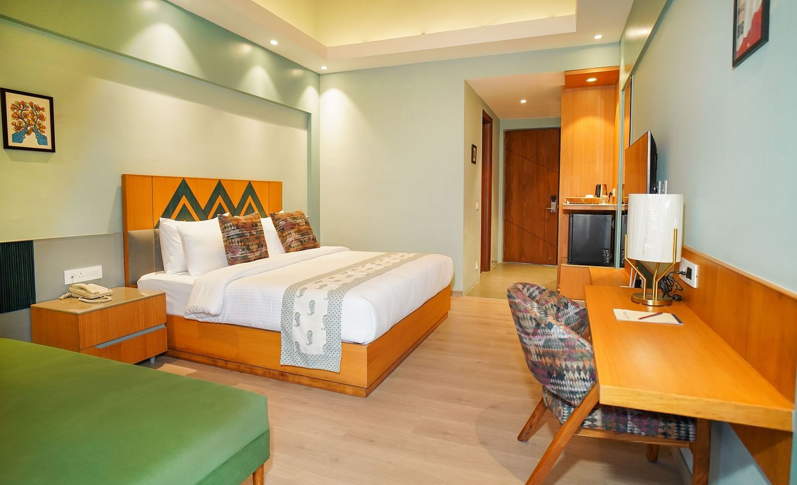 A brightly lit hotel room features a bed with white linens, a wooden headboard, a bedside table with a lamp, a desk, and a television, all in a warm-toned interior | Deluxe Room with Valley View | Sukama Resort & Spa, Nainital