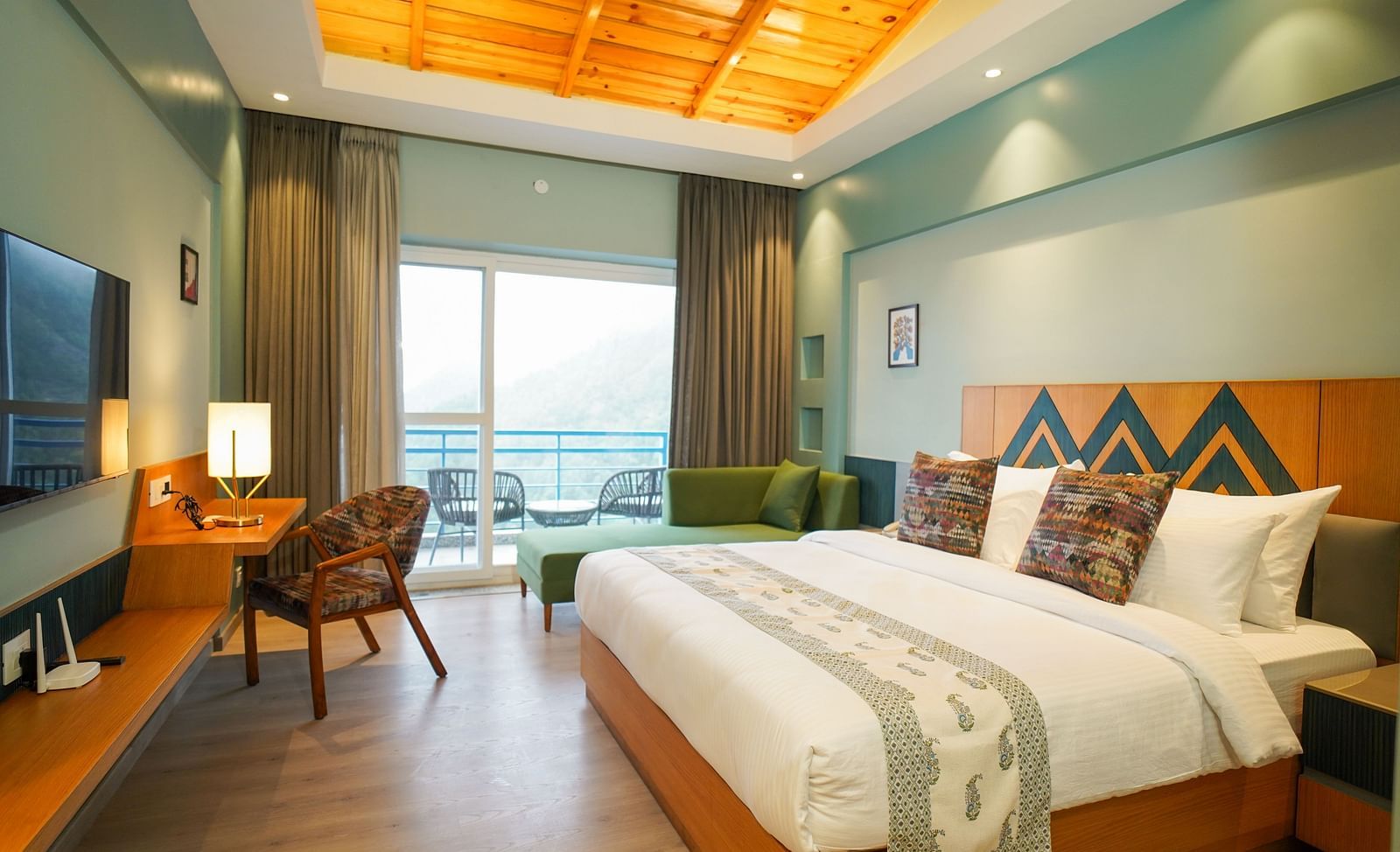 A spacious hotel room includes a large bed, a comfortable seating area with a sofa, and a balcony view of the hills | Deluxe Room with Valley View | Sukama Resort & Spa, Nainital