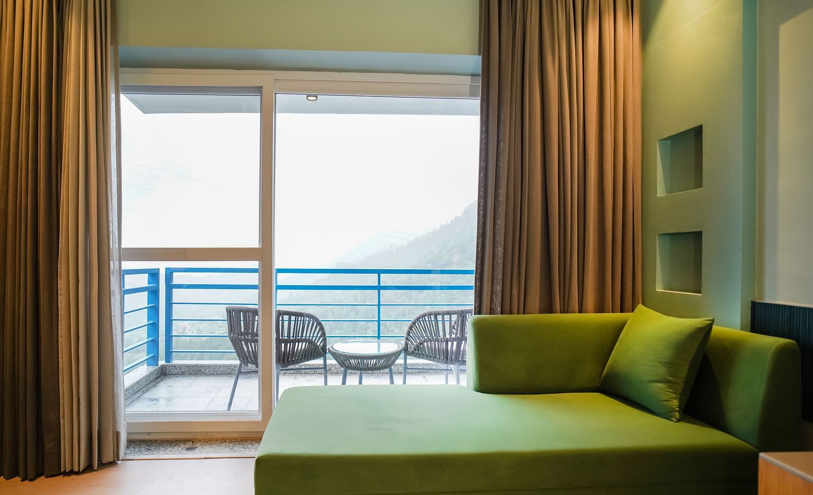A modern hotel room with a green sofa and a large sliding glass door opening to a balcony with outdoor seating and a view of the hills | Deluxe Room with Valley View | Sukama Resort & Spa, Nainital
