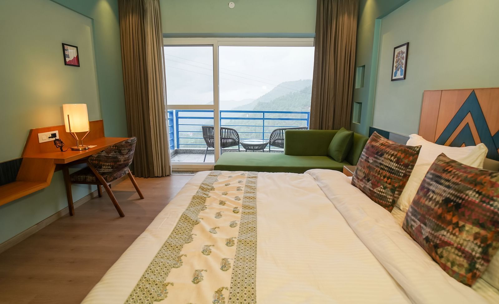 A bright hotel room features a large bed with decorative pillows, a desk and chair near a window, and a view of an outdoor area | Deluxe Room with Valley View | Sukama Resort & Spa, Nainital