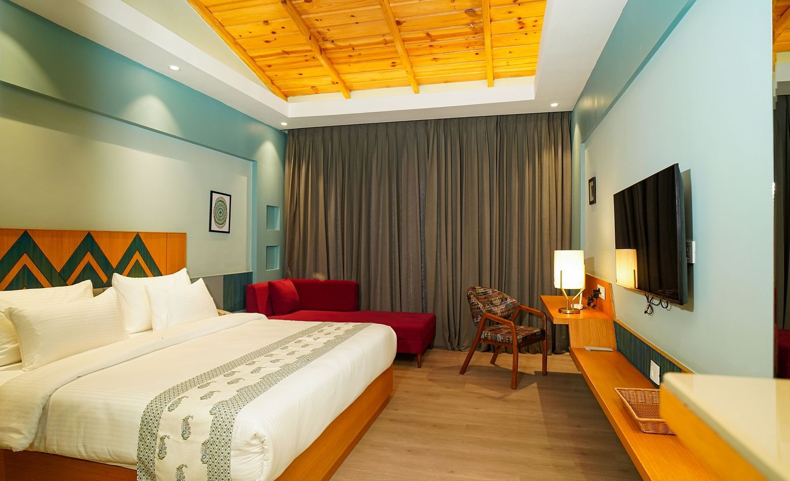A hotel room with a large bed featuring white linens and a wooden headboard also includes a wall-mounted television and a view of curtains and a patterned wall | Deluxe Room with Valley View | Sukama Resort & Spa, Nainital