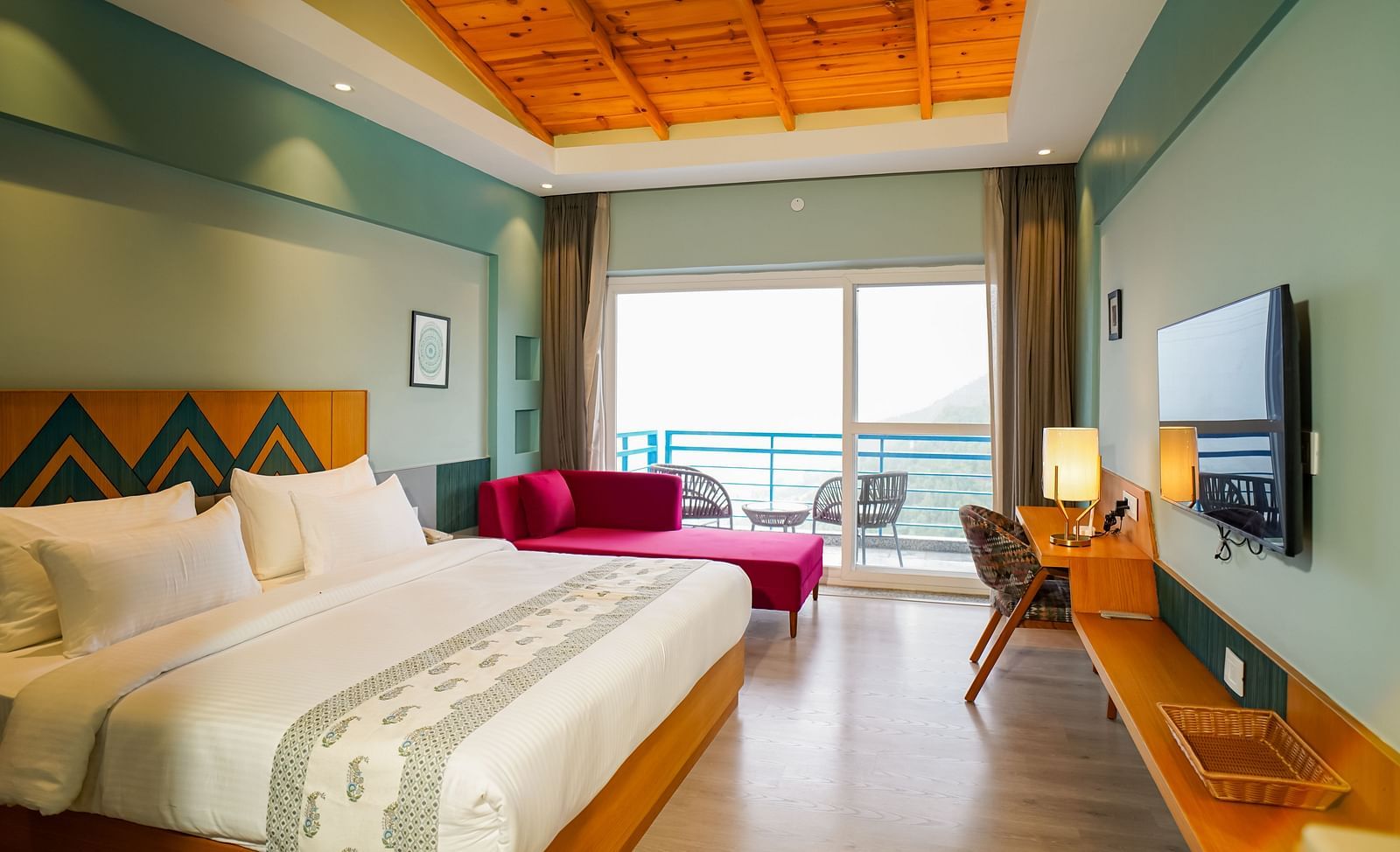A spacious hotel room includes a large bed with white bedding, a wooden headboard, and a purple chaise lounge at the foot, with a balcony visible through large glass doors | Deluxe Room with Valley View | Sukama Resort & Spa, Nainital