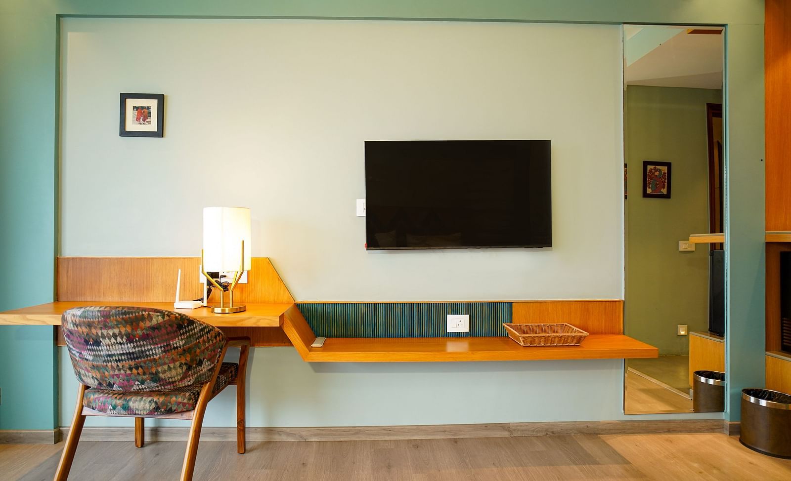 A hotel room showcases a large wall-mounted television above a long desk with a chair and lamp, complementing a bed with a wooden headboard and a white duvet | Deluxe Room with Valley View | Sukama Resort & Spa, Nainital