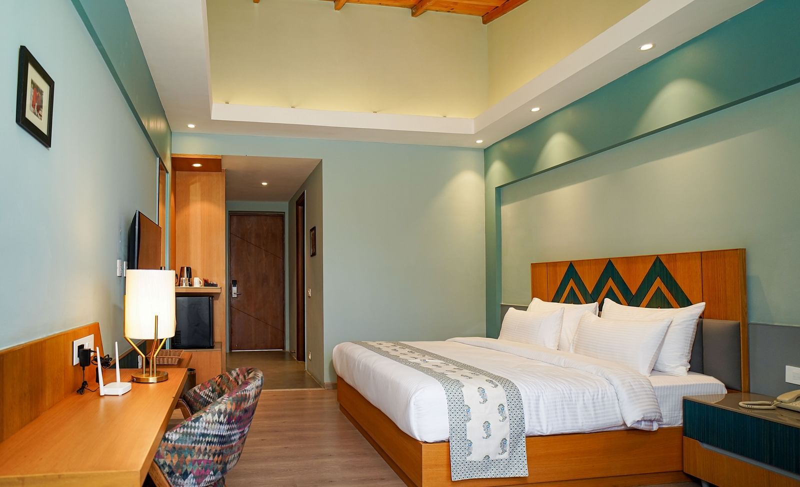 A well-lit hotel room features a bed with white linens, a wooden headboard, and bedside tables with lamps, alongside a desk area with a chair and a framed picture on the wall | Deluxe Room with Valley View | Sukama Resort & Spa, Nainital
