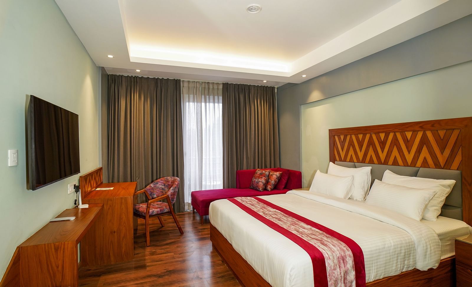 A hotel bedroom shows a neatly made bed with red accents, a sofa, and a television on the opposite wall | Deluxe Room with Himalayan View | Sukama Resort & Spa, Nainital