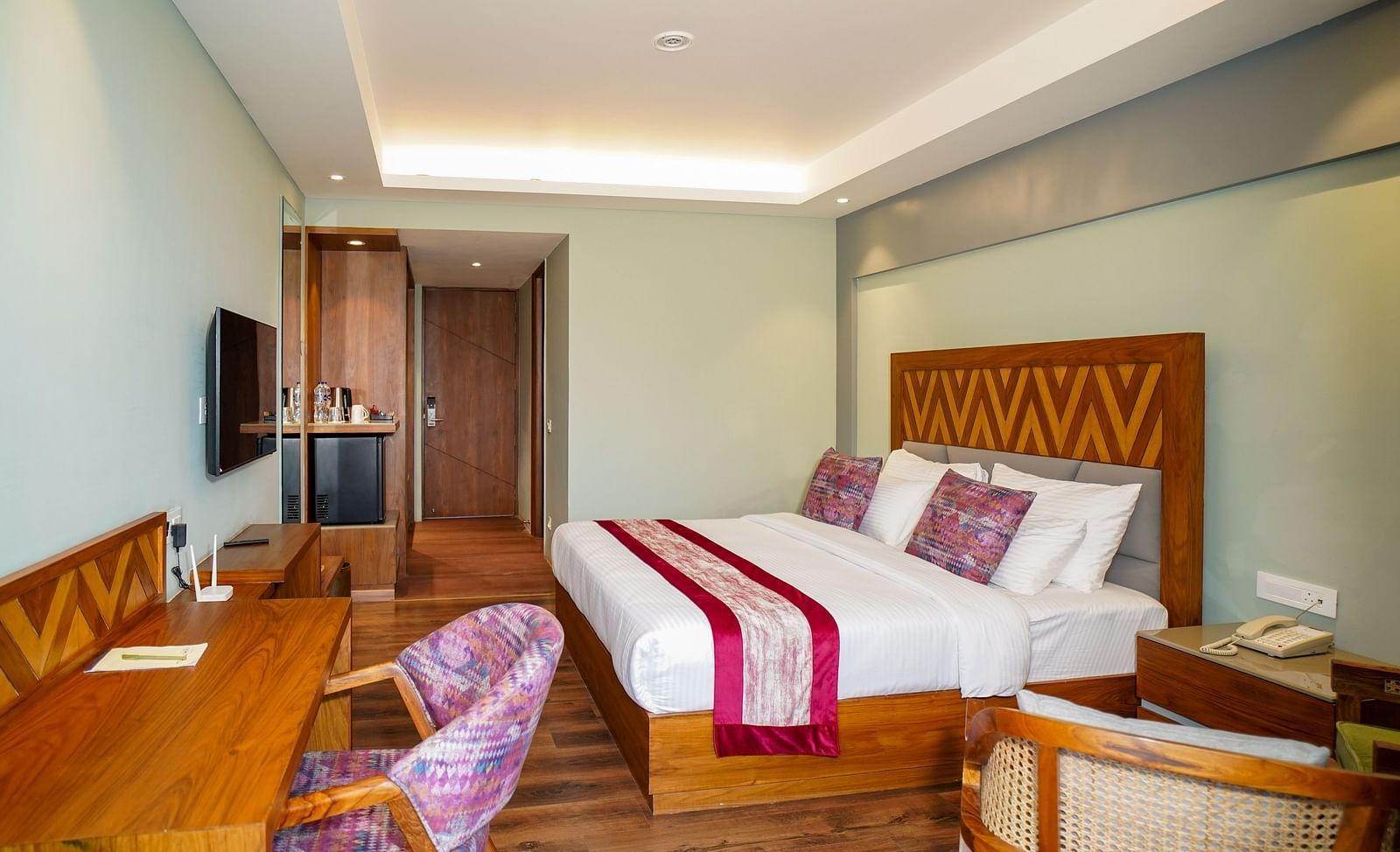 A hotel bedroom featuring a bed with white and red linens, and a seating area with chairs and a table | Deluxe Room with Himalayan View | Sukama Resort & Spa, Nainital