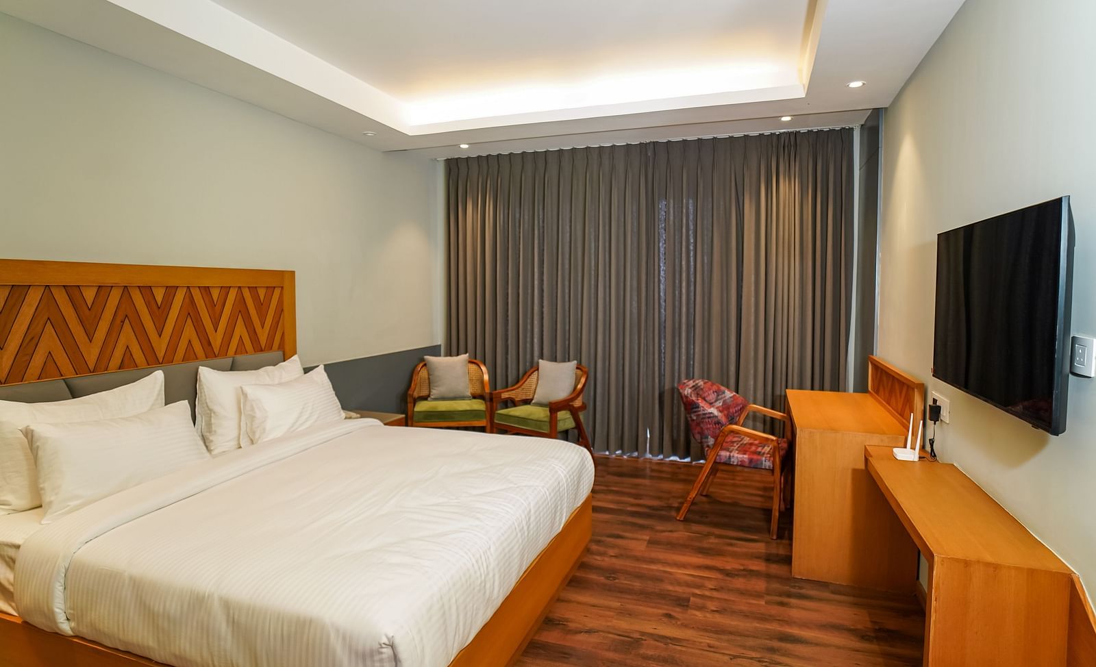 A spacious hotel room showcasing a large bed, a desk area with a chair, and a flat-screen TV on a stand | Deluxe Room | Sukama Resort & Spa, Nainital