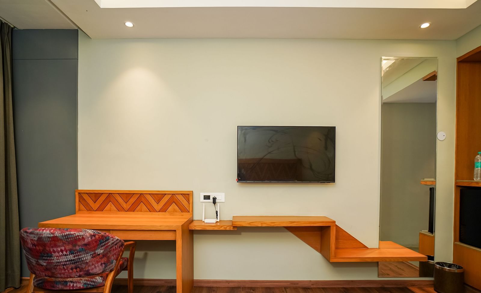 A modern hotel room with a long wooden desk and shelving unit, a chair, and a wall-mounted television | Deluxe Room | Sukama Resort & Spa, Nainital
