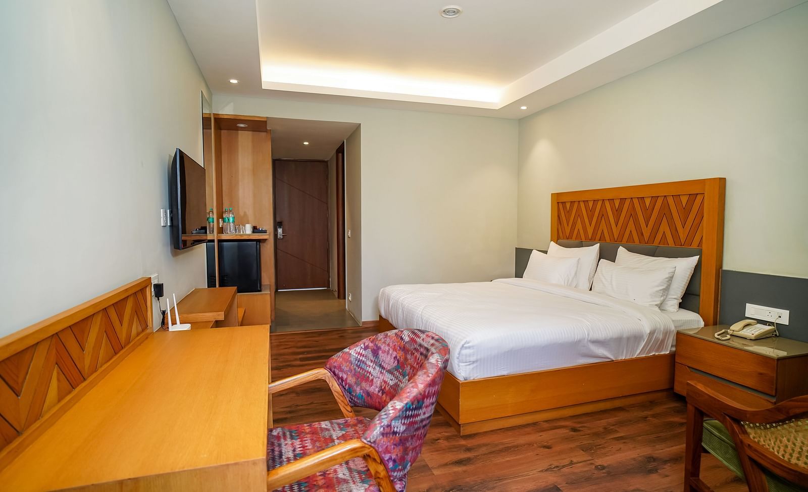 A bright hotel room featuring a large bed with a wooden headboard, a desk area, and a bedside table | Deluxe Room | Sukama Resort & Spa, Nainital