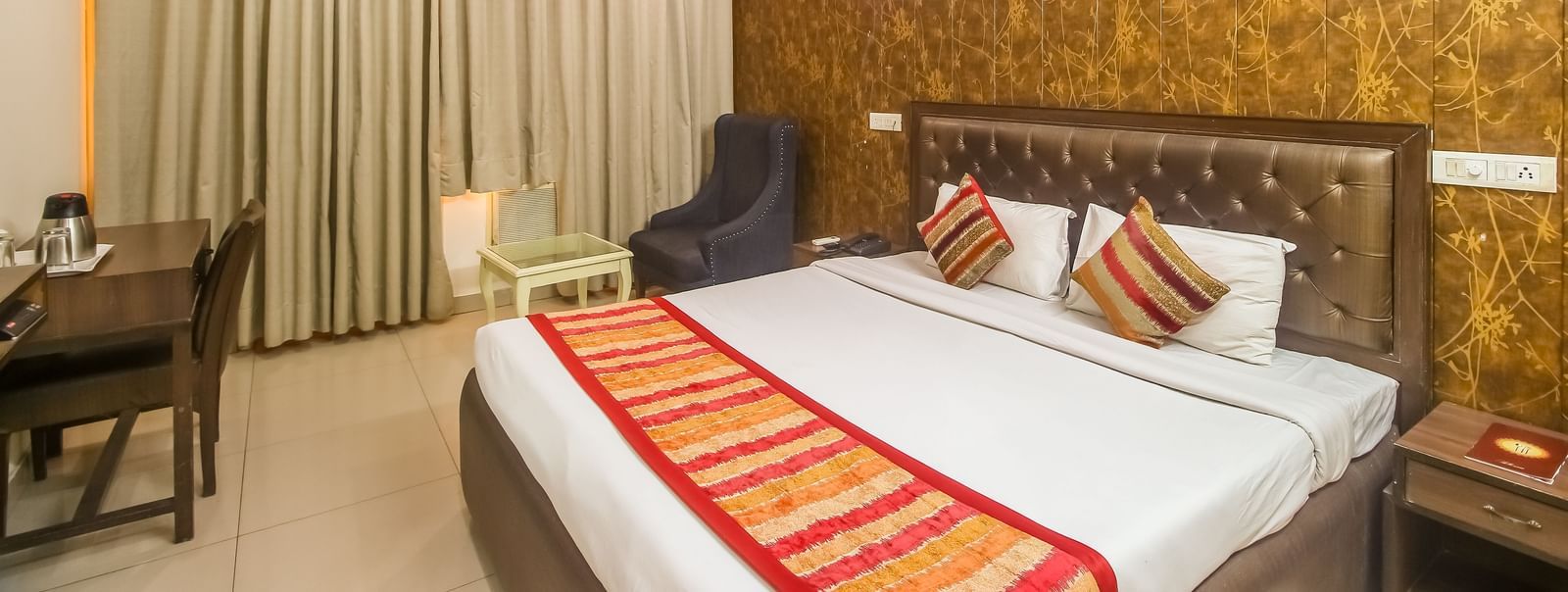 A hotel room with a textured accent wall, a plush bed with decorative pillows and a runner, a dark armchair, and a desk, illuminated by warm lighting. - Hotel Diamond Plaza, Chandigarh.