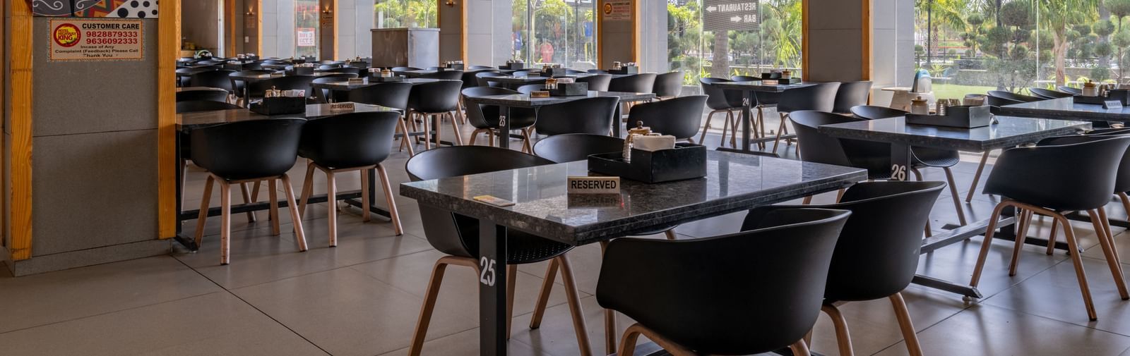 Dining area at Hotel Highway King, Bagru, with multiple tables and chairs, large windows, and wooden ceiling accents.