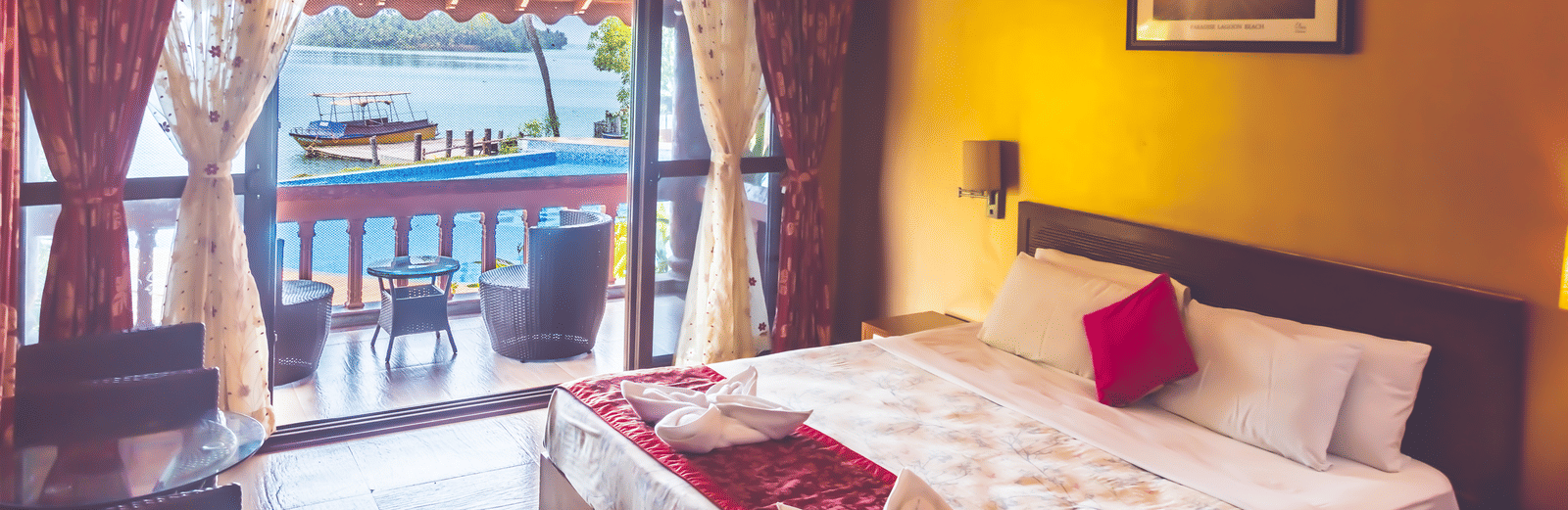 Room with a queen size bed, ceiling fan, framed picture, and view of the river through the window at Paradise Lagoon Resort, Udupi.