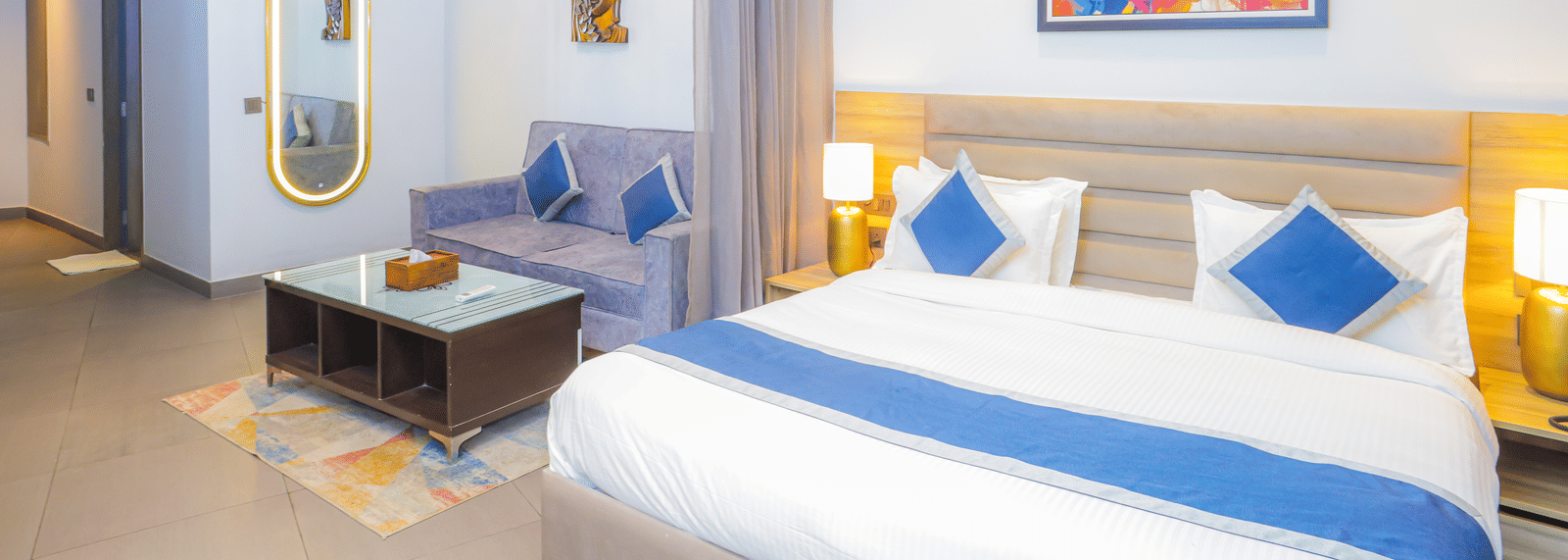 Bright bedroom with a king size bed, colourful wall art, and warm bedside lighting at Livbox Supernova, Noida.