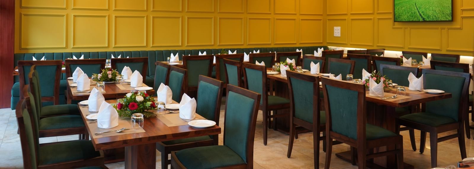 A wide shot of a dining hall with multiple tables set for a meal. The chairs are dark green, and the walls are yellow. A TV is visible on the far wall.