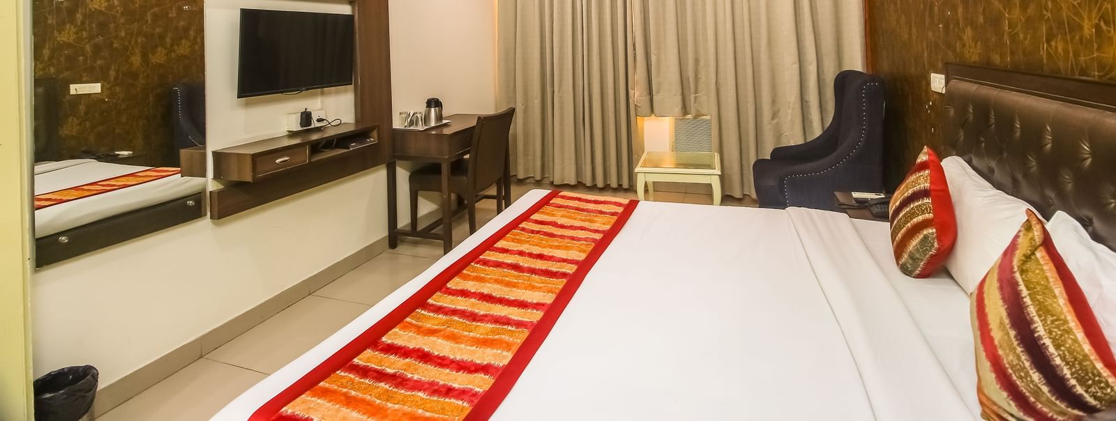 A hotel room with a plush bed featuring a patterned runner, a TV, a desk, a dark armchair, and a textured accent wall, lit by soft lighting. - Hotel Diamond Plaza, Chandigarh.
