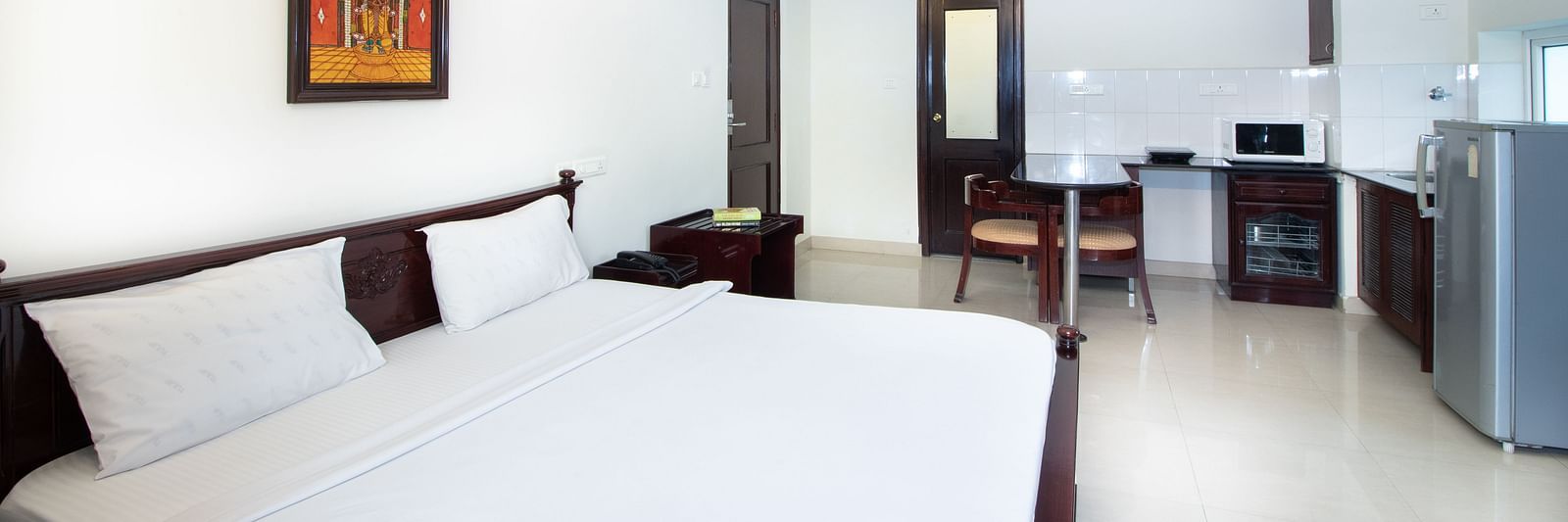 A room with a double bed featuring white linens, a ceiling fan, a framed picture on the wall, a dining counter, a refrigerator, and the kitchenette area at Sanctum Suites Indiranagar, Bangalore.