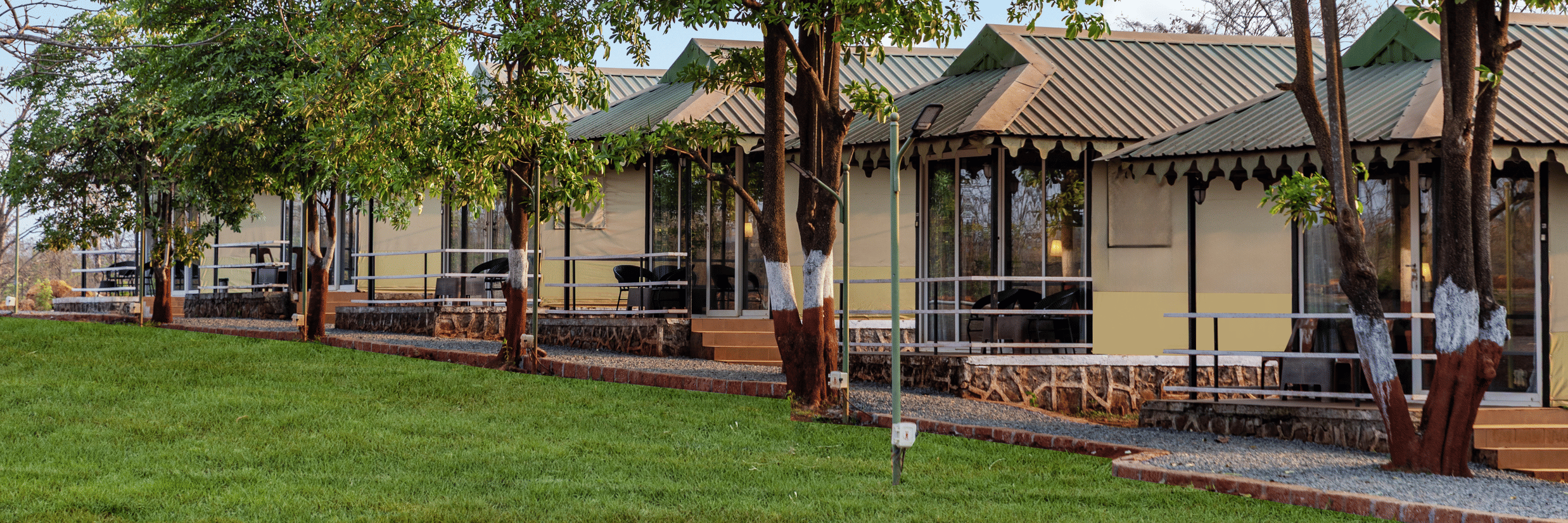 Nature Trails Kudalika Swiss-style cream cottages with green corrugated roofs, glass frontage and private decks, seen through a row of trees beside a green lawn.
