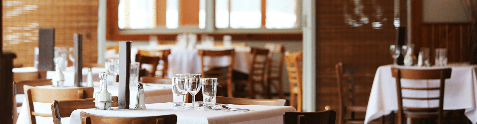 The bright interior of a restaurant with wooden tables and chairs, crisp white tablecloths, and bamboo window blinds.