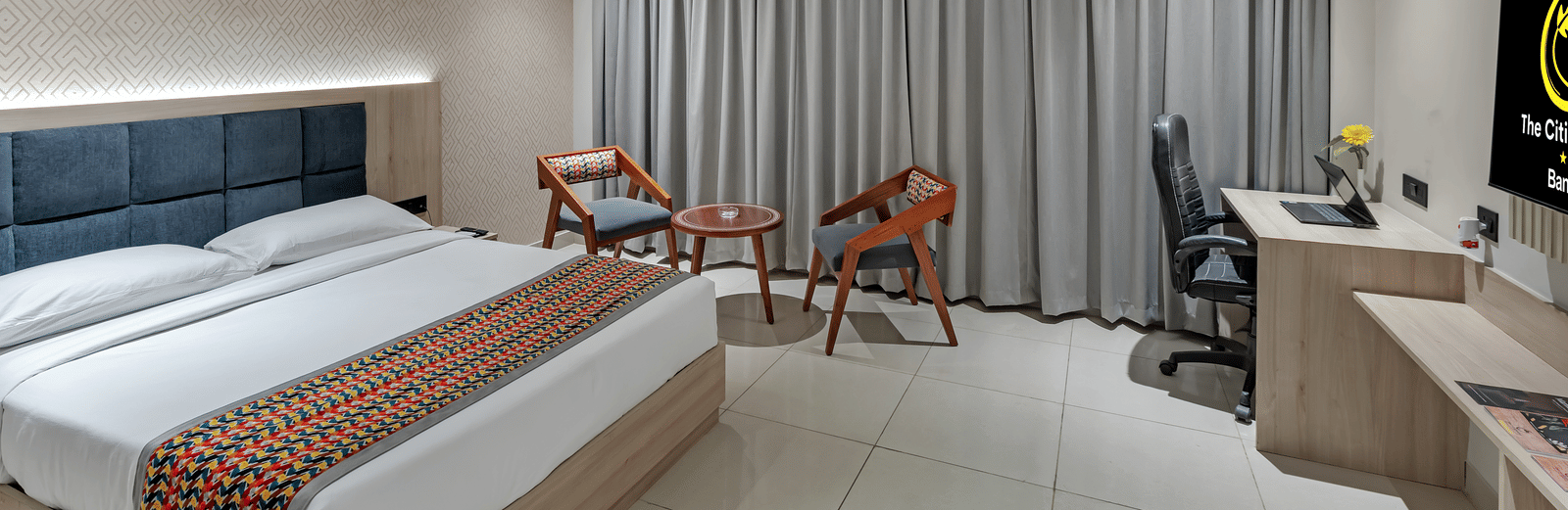 A well-lit hotel room with a queen size bed featuring a colourful patterned throw, a seating area with two chairs and a small table, a work desk with a chair, and a television | The Citi Residenci Hotel Banskopa