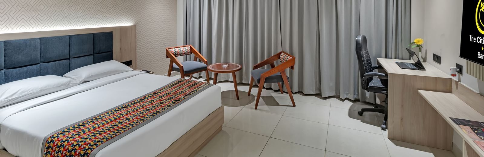 A well-lit hotel room with a king size bed featuring a colourful patterned throw, a seating area with two chairs and a small table, a work desk with a chair, and a television | The Citi Residenci Hotel Banskopa