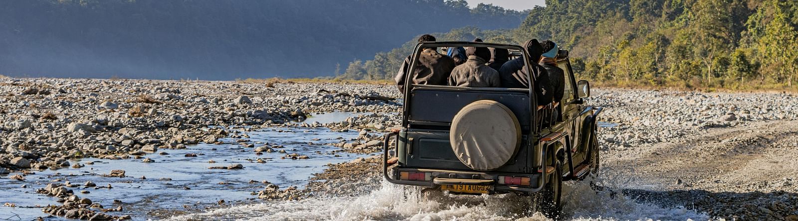 A jeep full of people drives through a shallow, rocky riverbed in Jim Corbett National Park, surrounded by forested hills.
