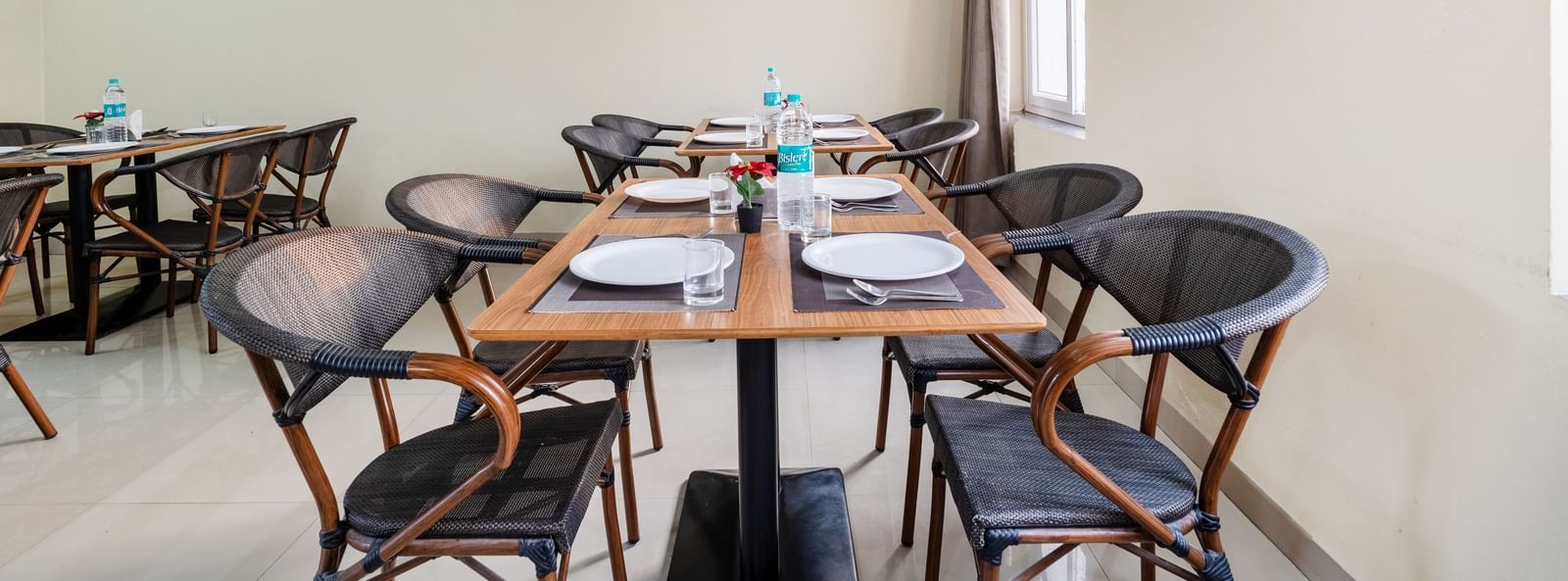 An empty dining area at Destino by Monday Hotels, Gachibowli, with 2 dining tables and woven chairs, set up with plates and water bottles.