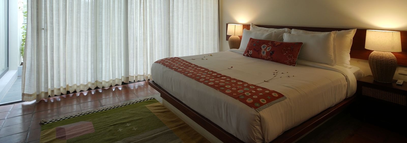 A deluxe room at Vaasta Vagamon featuring a large bed with decorative throw pillows and a patterned runner, and long drapes.
