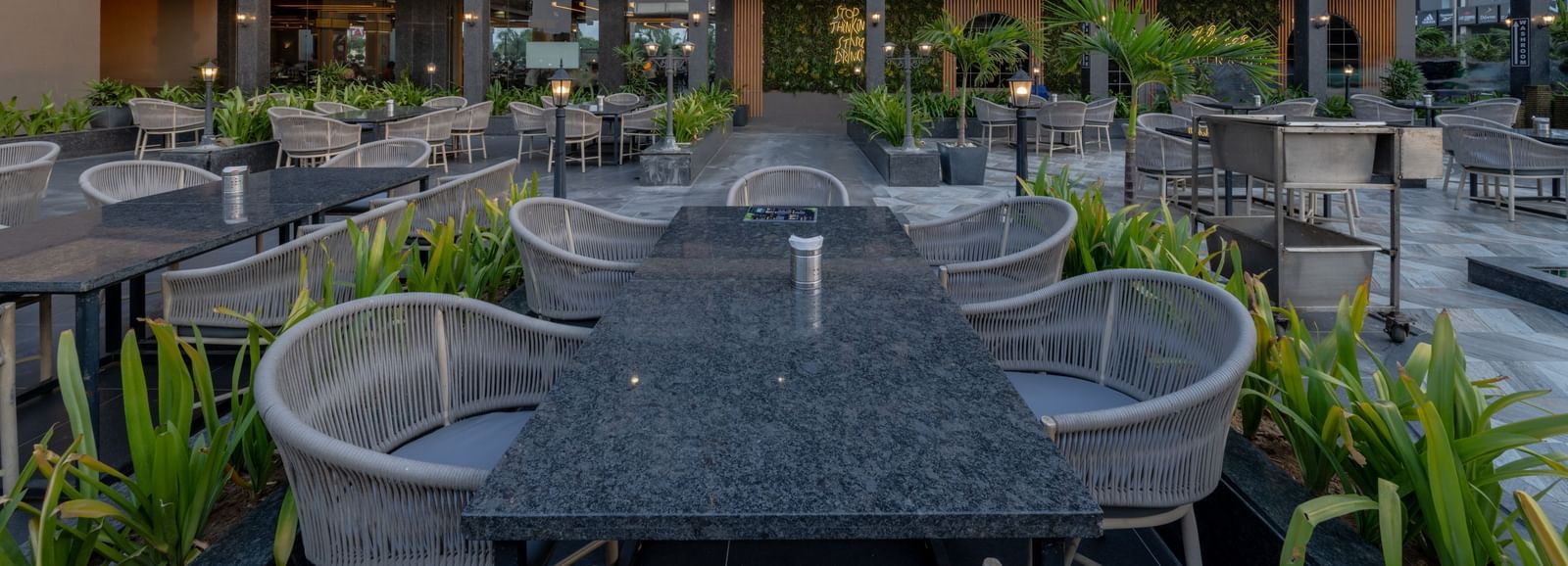 Outdoor dining area at Hotel Highway King, Bagru, with tables, chairs, plants, and multi-storey hotel building in the background.