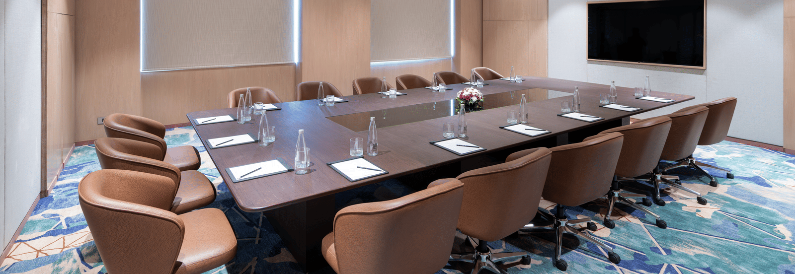 Diagonal view of a conference room featuring a projector, large table, and cushioned chairs at Merlis Hotel, Coimbatore.
