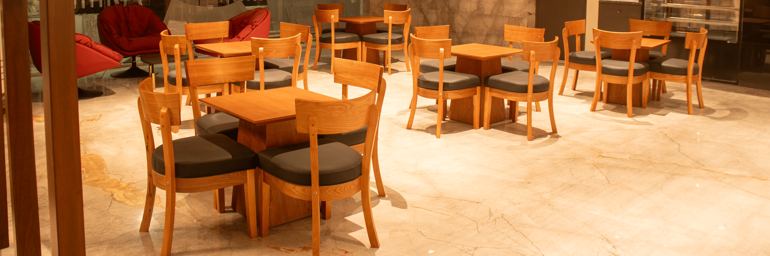 A hotel lobby area with wooden tables and chairs,  cushioned chairs, and ceiling lights arranged in a grid pattern.