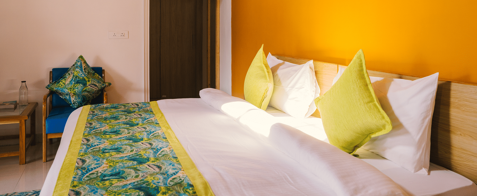 A side view of a neatly made bed with a photo frame above the headboard, two night lamps on either side, and a door in the background at BluSalzz Villas - The Green Roof, Bhimtal.