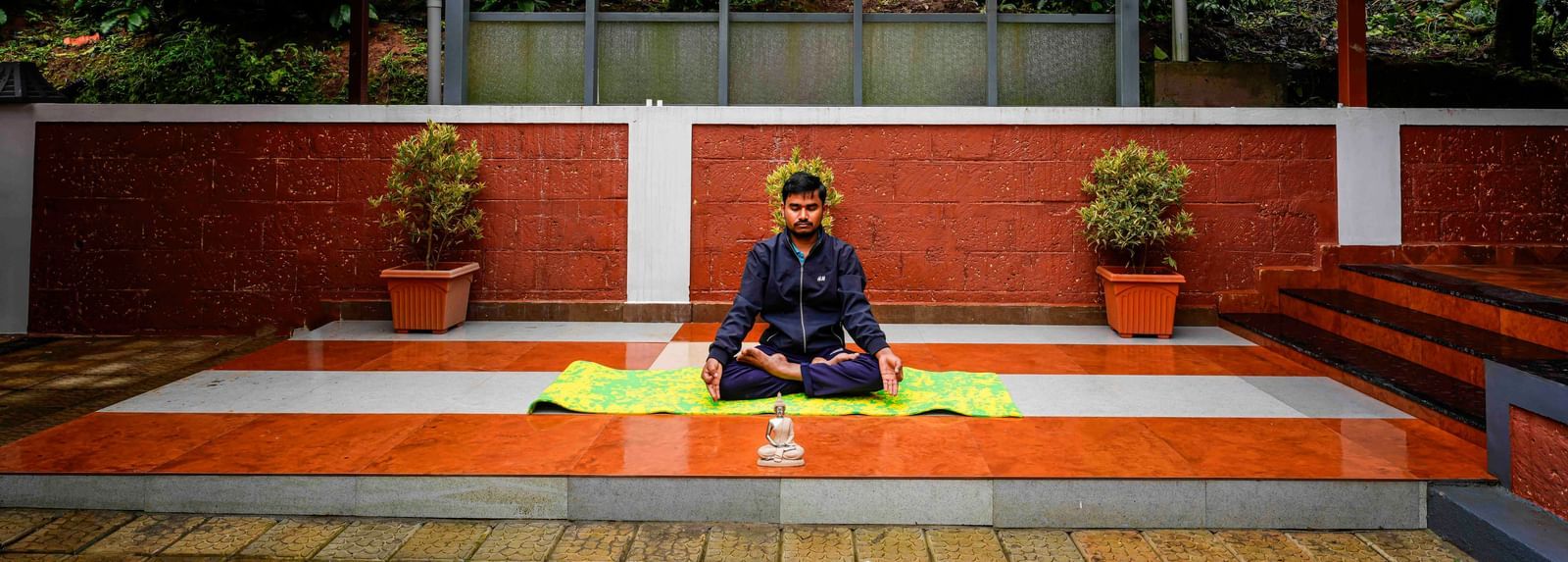An individual is meditating in a serene, partially covered outdoor yoga space at Bhuthanakadu Retreat, surrounded by lush greenery and red-tiled walls.