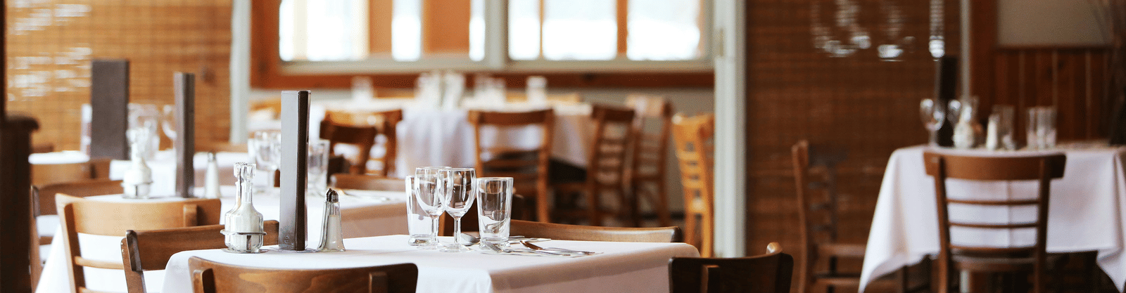 The bright interior of a restaurant with wooden tables and chairs, crisp white tablecloths, and bamboo window blinds.