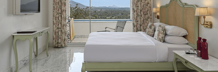 Side view of a  well-lit Plunge Pool Suite at Khas Mahal Suites, Udaipur featuring a bed, seating area, and a large window with a view.