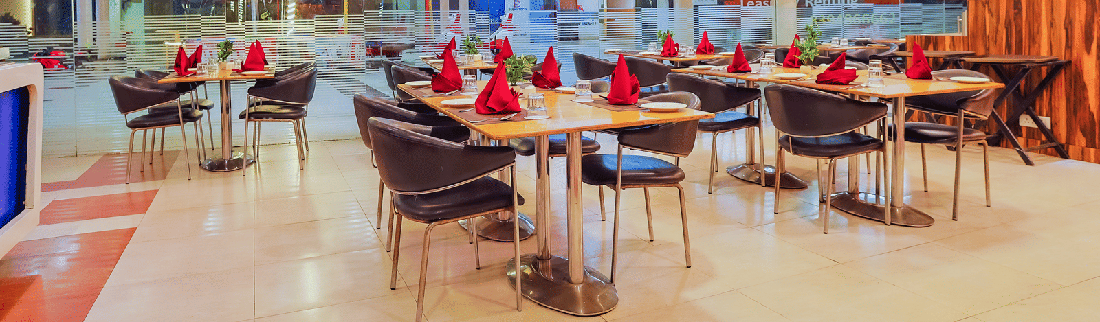 A bright and open restaurant dining area with several tables and chairs, a buffet or serving station in the background, and large windows.