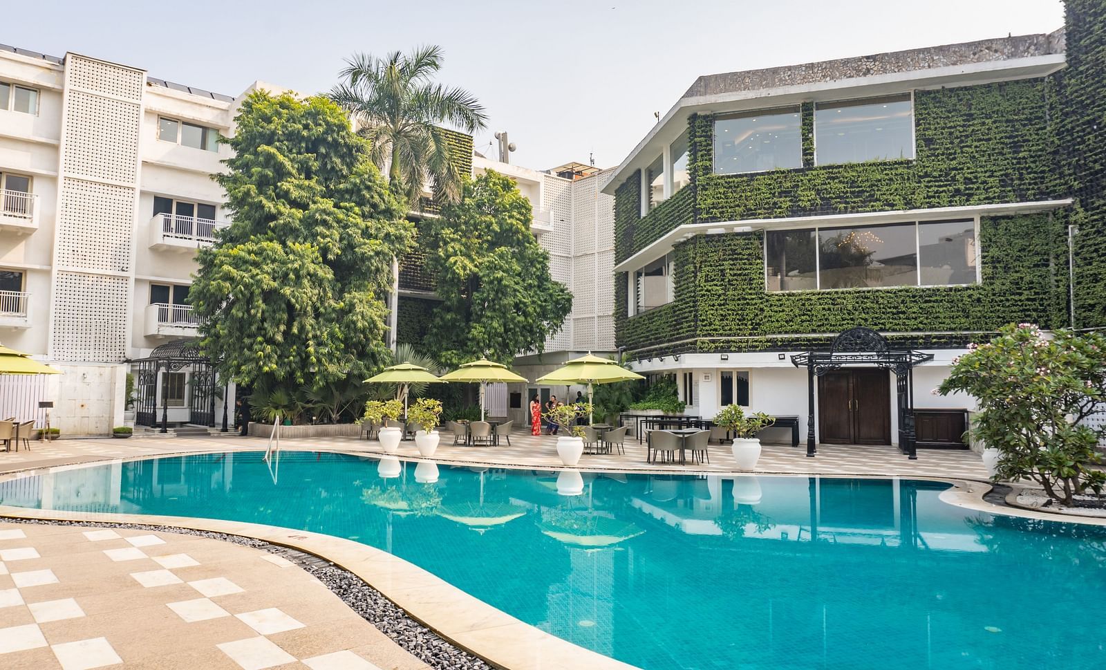 Outdoor swimming pool surrounded by a tiled deck, sun loungers with green umbrellas, and multi-storey buildings, including one with a lush green vertical garden.
