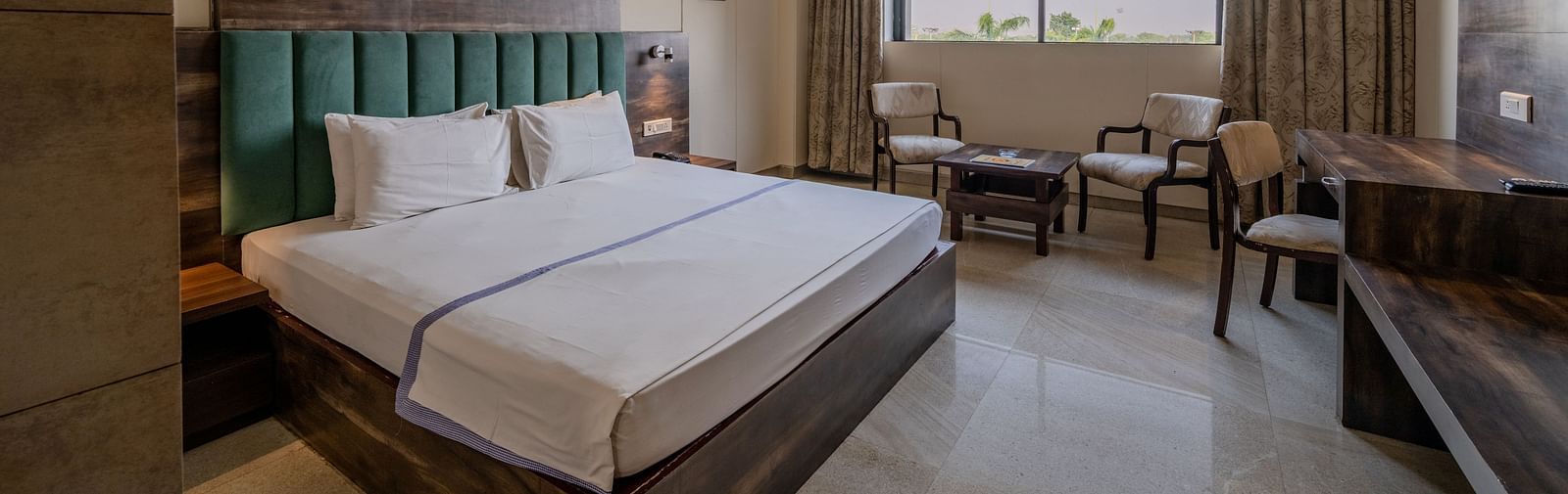 An accommodation in Hotel Highway King, Behror, featuring a plush double bed with upholstered headboard, seating area for two, a work desk and glass windows with open curtains.