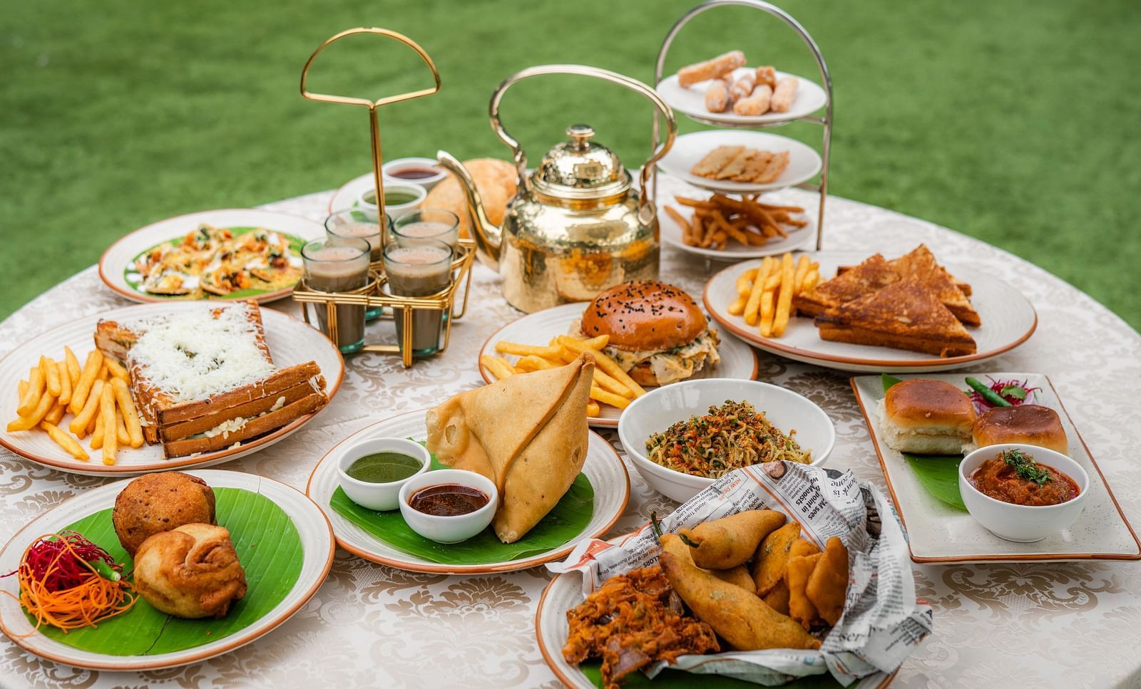 A breakfast table where varieties of savouries served in each plat consisting of samosa, french fries, sandwiches, salsa, fritters, bun, etc. There is also a kettle and hot tea served in four cups. 