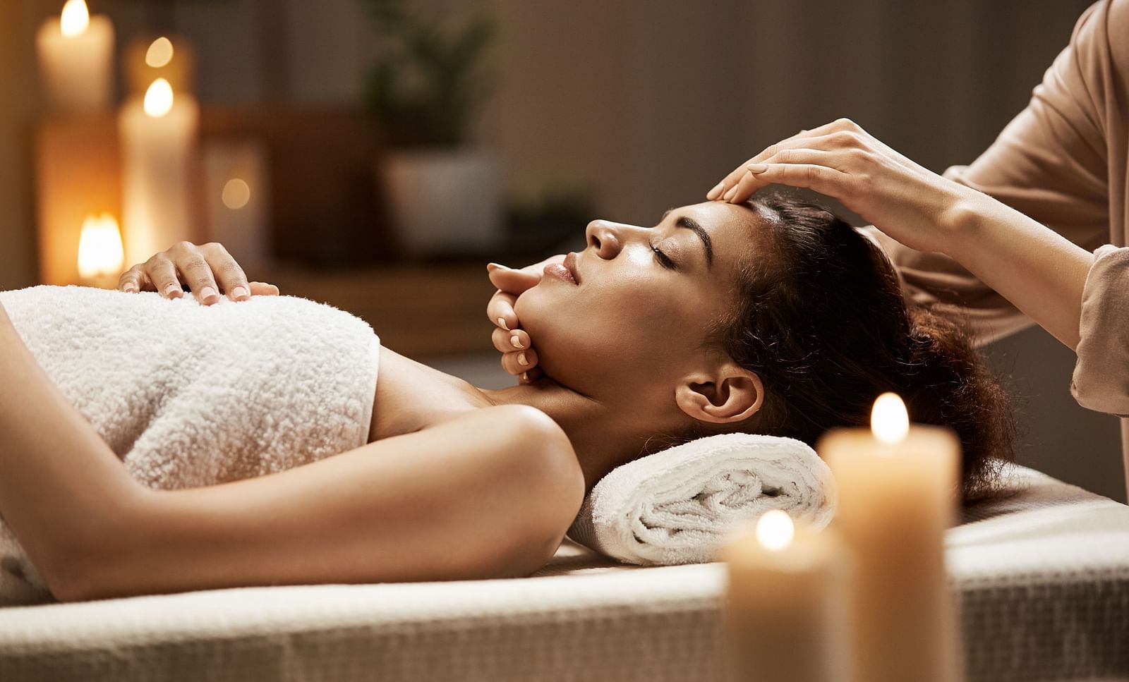A woman lies on a massage table, draped in a white towel, receiving a head massage in a softly lit, tranquil spa setting with candles.