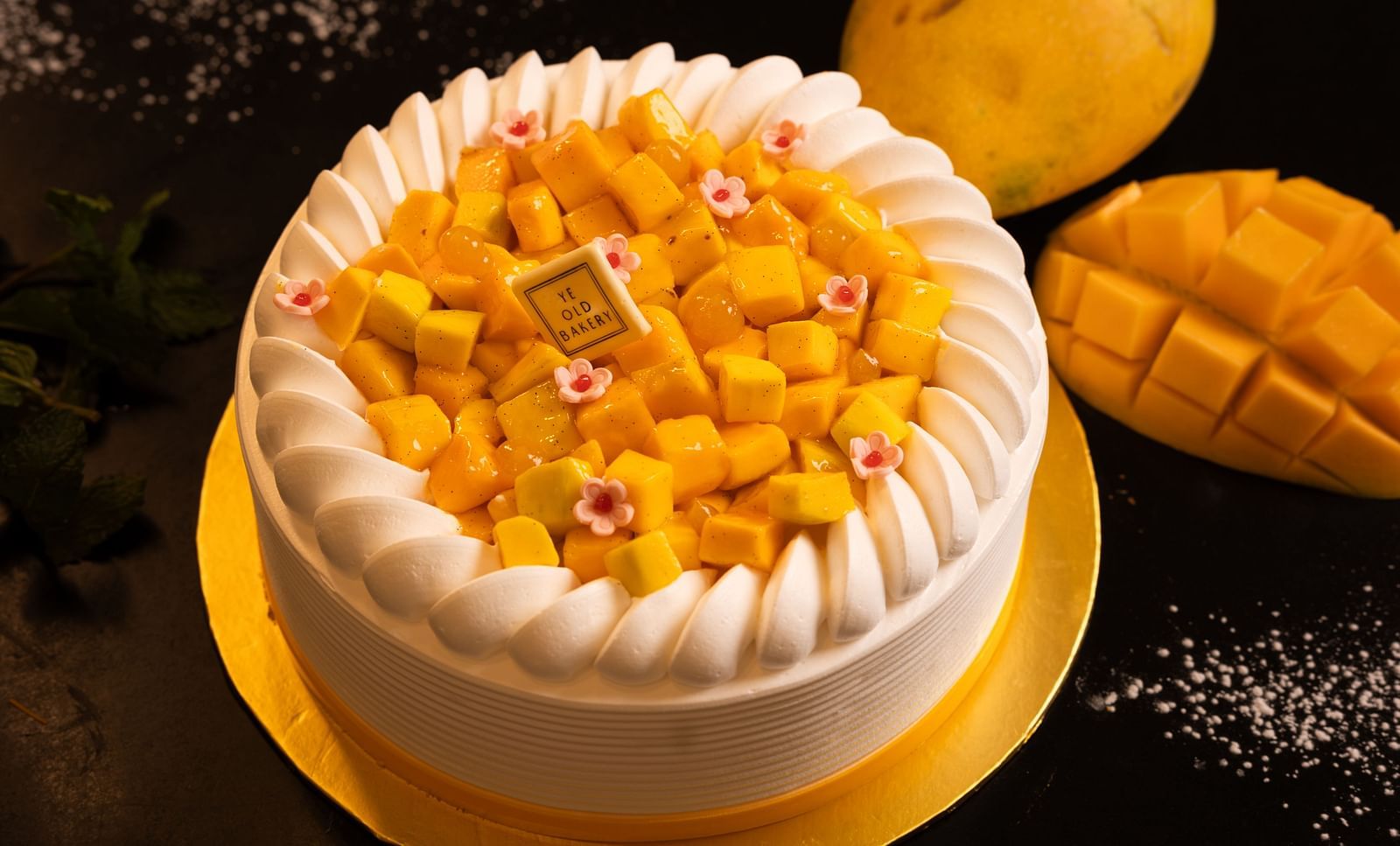 A round mango cake decorated with whipped cream along the edges and topped with diced mango pieces and small pink flowers, placed on a golden cake board with whole and sliced mangoes in the background.