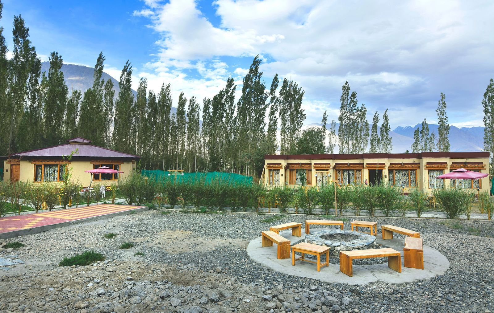 An overview of White Sands Resort, Ladakh featuring a communal seating area with wooden benches and a fire pit surrounded by gravel, with cottages and trees in the background under a cloudy sky.
