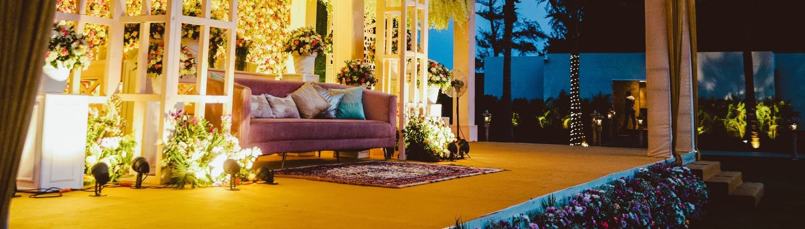 A side angle shot of a mandap in the lawn of Kaldan Samudhra Palace featuring a chandelier, a well-decorated sofa and ornamental plants hanging from the top
