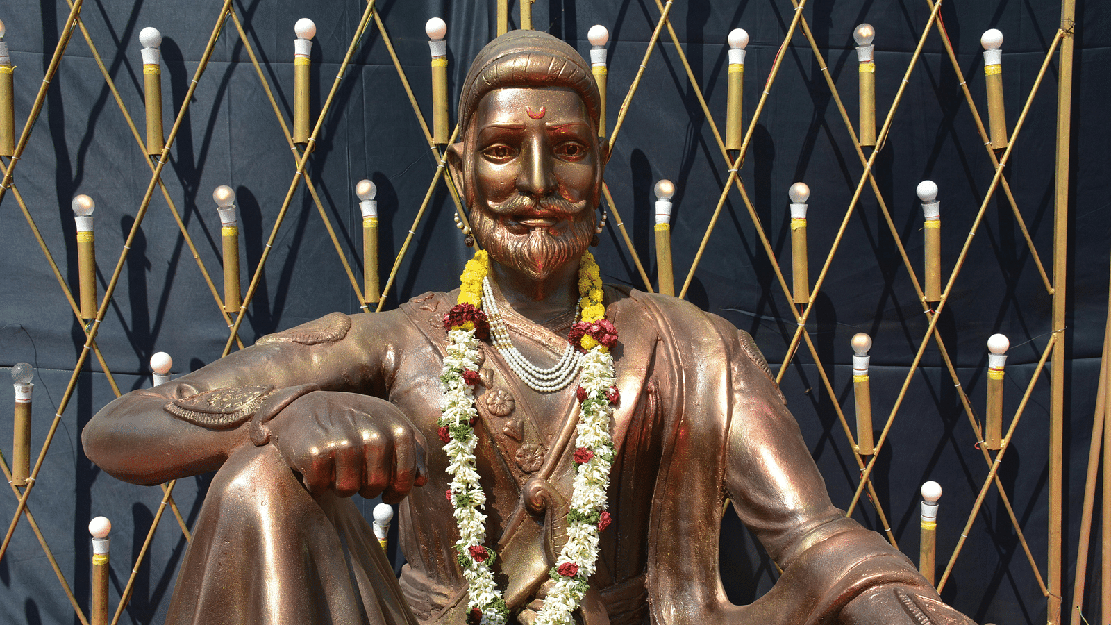 Bronze statue of Chattrapati Shivaji Maharaj sitting magnificently, decorated with garlands and flower decors.