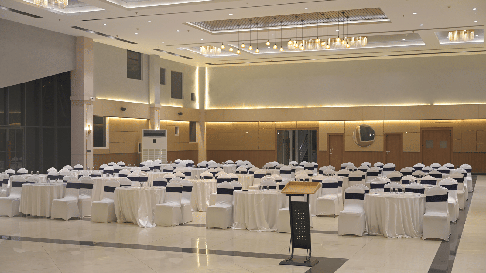 A front-angled view of the banquet hall set with round tables and white seating under warm ceiling lights at Hotel Sonar Bangla Mayapur.